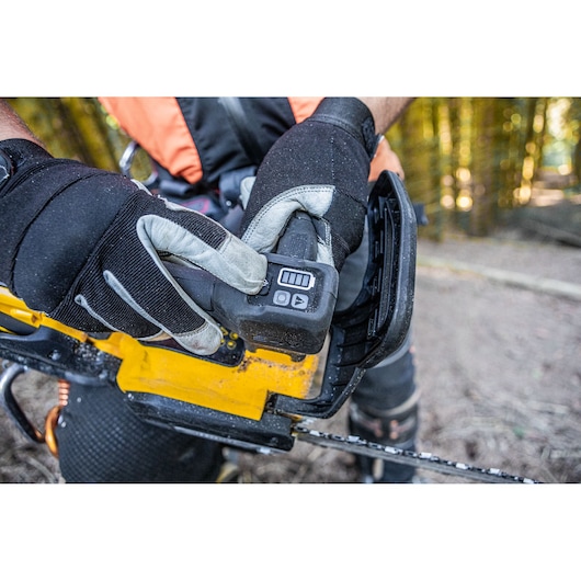 Close-up of a person wearing protective gloves and an orange vest, inserting a battery into a yellow DEWALT chainsaw outdoors in a forest setting.