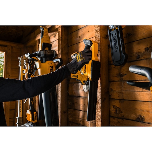 A person wearing black gloves is placing a yellow and black DEWALT power tool, specifically a hedge trimmer, onto a wooden wall rack in a workshop. Other DEWALT tools are visible hanging on the wall nearby.