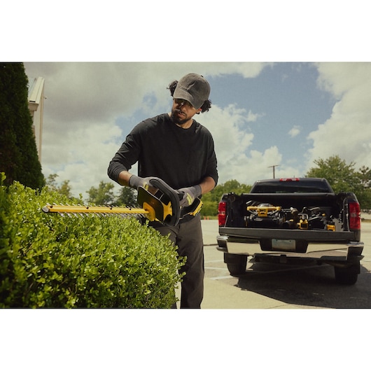 Person trims hedge with DEWALT cordless hedge trimmer, pickup truck with tools in background outdoors.
