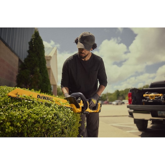 Person trims hedge with DEWALT cordless hedge trimmer outdoors near a parked truck on a sunny day.