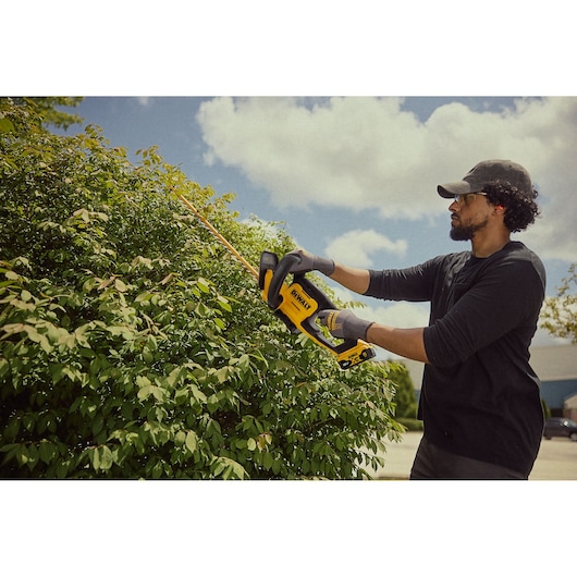 Person trims bushes using a DEWALT cordless hedge trimmer outdoors on a sunny day.