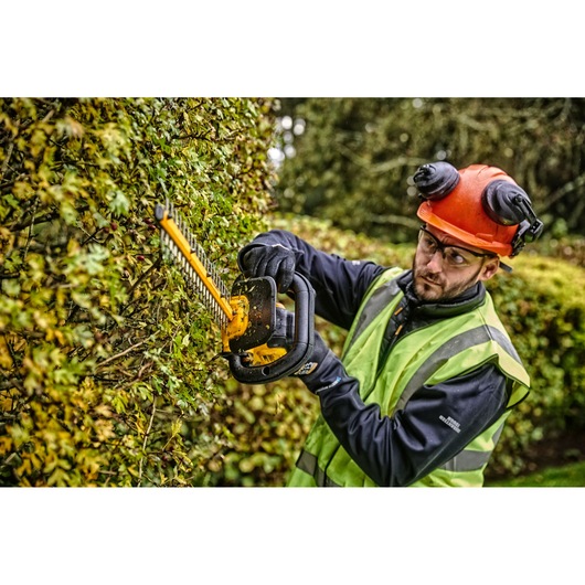 A person wearing safety gear, including an orange helmet and a reflective vest, uses a DEWALT DCMHT563N hedge trimmer to cut hedges outdoors. The hedge trimmer is yellow and black and designed for professional gardening and landscape maintenance.