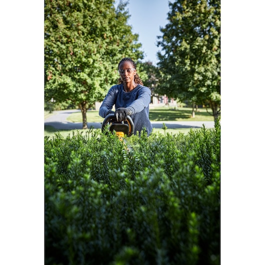 A person wearing gloves uses a DEWALT hedge trimmer to trim green bushes outdoors, with trees and a clear sky in the background.