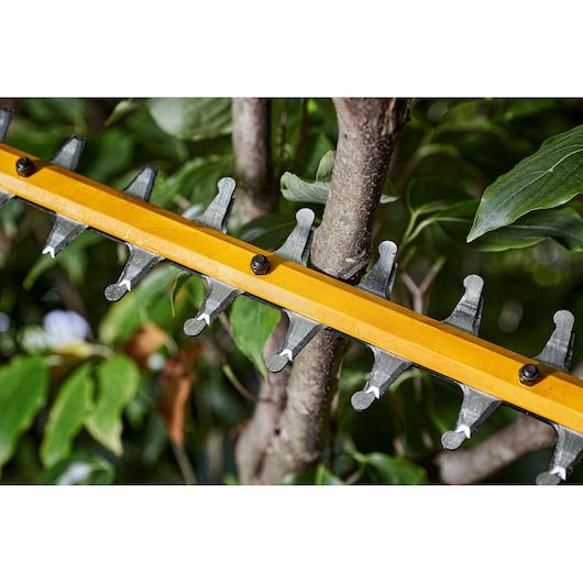 Close-up image of a yellow DEWALT hedge trimmer blade cutting through a tree branch, surrounded by green leaves.