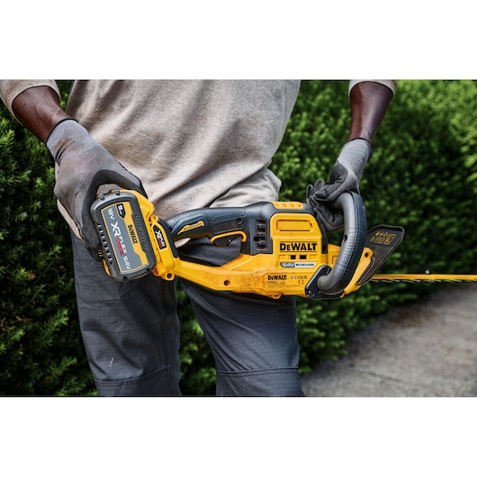 A person wearing gloves holds a DEWALT cordless hedge trimmer with a 54V XR FLEXVOLT lithium-ion battery, standing in front of a green hedge.
