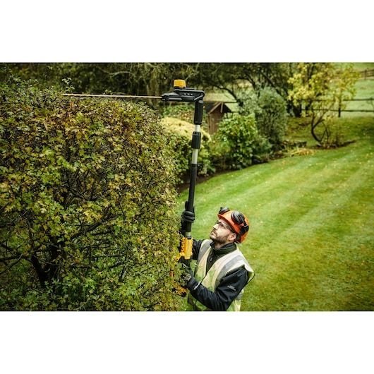 A person using a DEWALT pole hedge trimmer (model DCMPH566P1) to trim a large hedge in a garden. The person is wearing protective gear, including gloves, a helmet, and a reflective vest.
