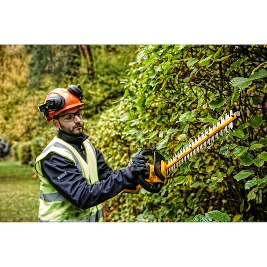 A person wearing safety gear and a helmet is using a DEWALT cordless hedge trimmer to trim green bushes outdoors.