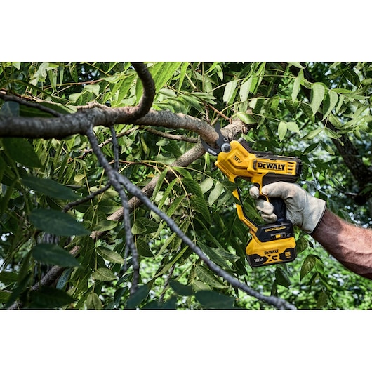A person wearing gloves uses a yellow DEWALT 18V cordless pruning shears to cut a thick tree branch surrounded by green leaves.