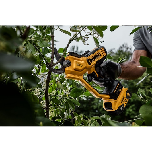 A person wearing a glove is using a yellow DEWALT 18V cordless battery-powered pruning shear to cut a tree branch outdoors among green leaves.