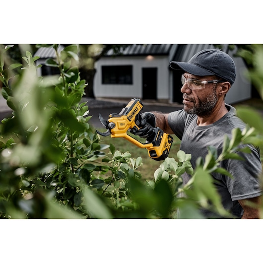 A person using a DEWALT 18V cordless electric pruning shear (DCMPP568P1) to trim branches on a shrub outdoors. The person is wearing a cap and gloves. The focus is on the tool and the pruning activity, with greenery in the foreground and a building in the background.