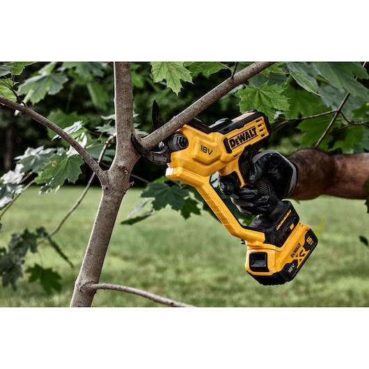 A person wearing gloves uses a DEWALT 18V cordless pruning shear to cut a branch from a tree outdoors.