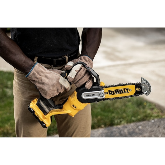 A person wearing work gloves and beige pants is holding a DEWALT cordless chainsaw outdoors. The DEWALT logo and chain are clearly visible.
