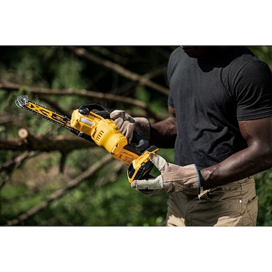 A person wearing work gloves is holding a yellow DEWALT cordless chainsaw outdoors, preparing to use it, with fallen branches and greenery in the background.