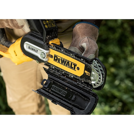 A close-up view of a person wearing gloves and holding a DEWALT cordless chainsaw, with the chain cover open to show the chain and bar.