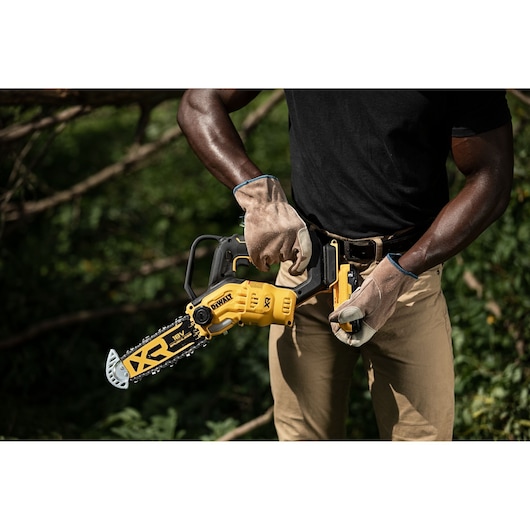 A person wearing gloves and work clothes is holding a DEWALT cordless pruning chainsaw outdoors, preparing it for use.
