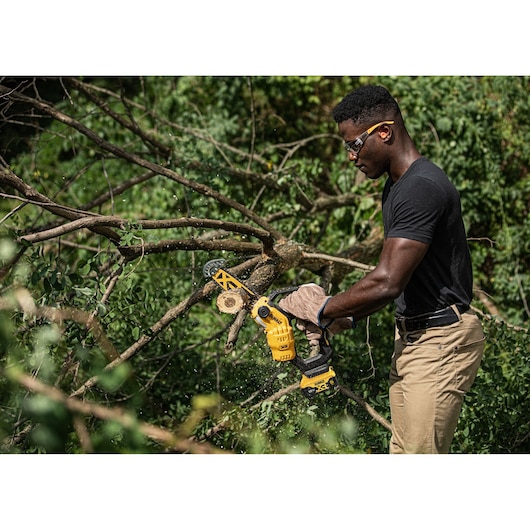 A person wearing protective gloves uses a yellow DEWALT DCMPS520P1 cordless chainsaw to cut tree branches outdoors.