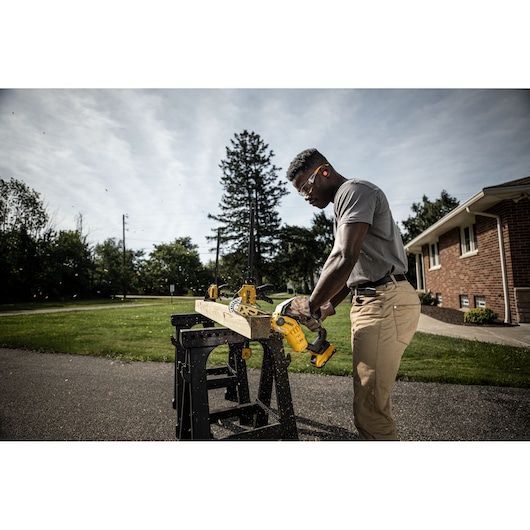 A person using a DEWALT DCMPS520P1 cordless chainsaw to cut a wooden plank outdoors, with the plank supported on a black workbench. The scene is set in a residential area with green grass and trees in the background.