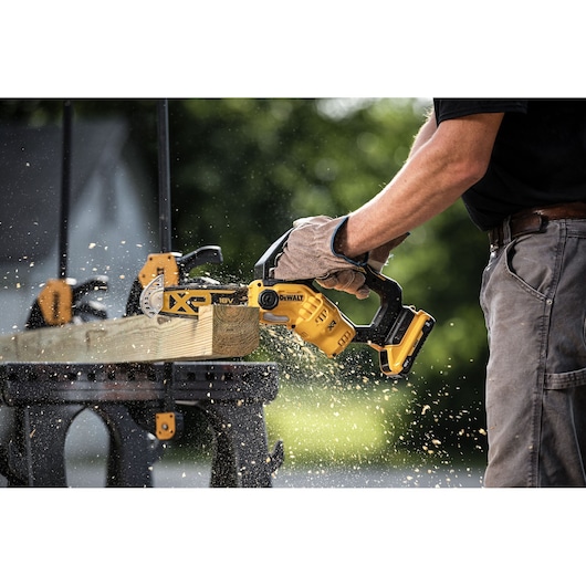 A person wearing gloves is using a DEWALT DCMPS520P1 cordless chainsaw to cut through a piece of wood placed on a workbench, with wood chips flying around.