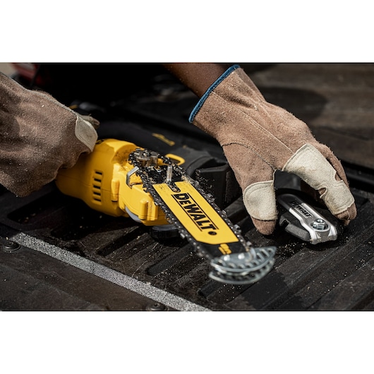A person wearing work gloves is adjusting a DEWALT yellow cordless chainsaw, model DCMPS520P1, on a black surface. The chainsaw has visible sawdust on it and the person is holding a tool.