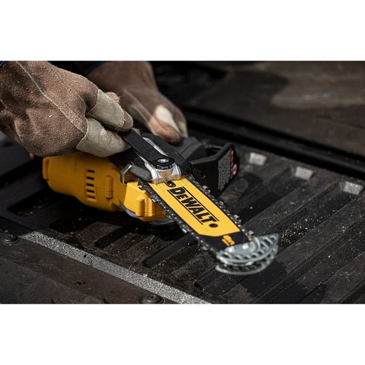 A close-up of gloved hands adjusting the chain on a DEWALT cordless chainsaw. The chainsaw is placed on a textured surface and the yellow DEWALT branding is visible on the blade.
