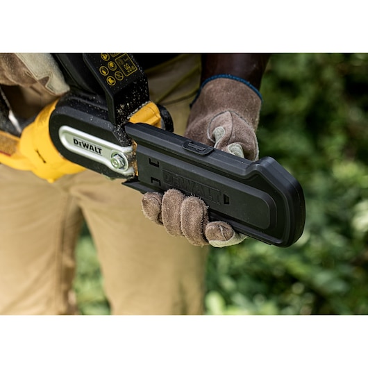 A person wearing work gloves holds a DEWALT battery-powered pole saw with a protective blade cover outdoors, surrounded by greenery.