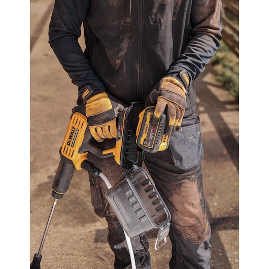 A person wearing work gloves and dirty work clothes is holding a DEWALT cordless pressure washer with attached battery and water hose.