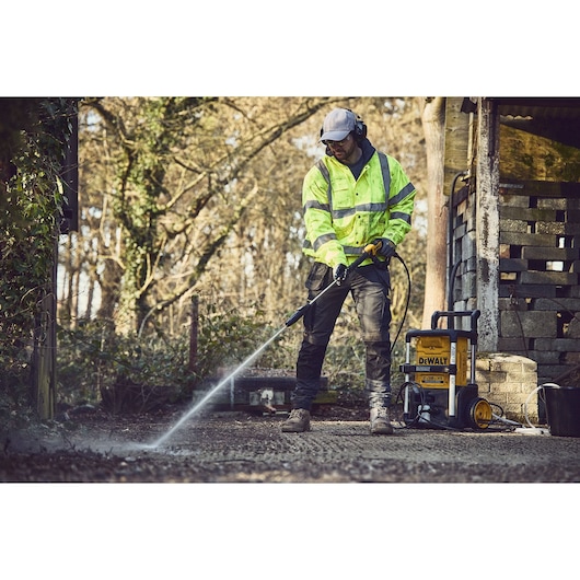 Person uses a DEWALT pressure washer outdoors on a paved surface beside a shed, wearing safety gear.