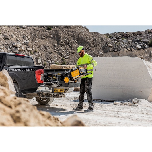 A construction worker wearing a neon yellow safety jacket and cap is unloading a DEWALT pressure washer from the bed of a black pickup truck at a quarry site. The surrounding area is filled with large rocks and stone blocks.