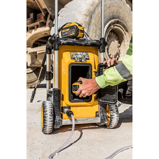 A close-up view of a DEWALT portable equipment with wheels, being operated outdoors. A person is placing or removing a battery from its compartment. The equipment is yellow and black, and it is connected to a hose.