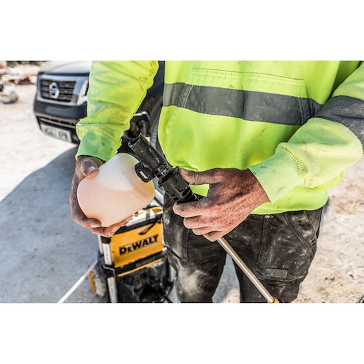 A person in a high-visibility jacket is attaching a soap dispenser to a DEWALT pressure washer outdoors, with a vehicle visible in the background.