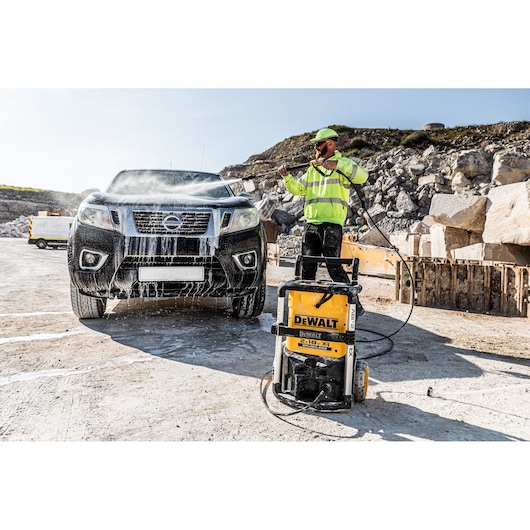 A person using a DEWALT pressure washer (model DCMPW1600) to clean a black pickup truck with water and soap outdoors at a worksite with rocks and construction materials in the background.