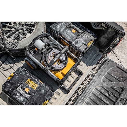 A DEWALT portable power washer and durable tool storage boxes are shown in the back of a vehicle, with a coiled hose visible. The equipment is dusty, indicating use in a rugged outdoor environment.
