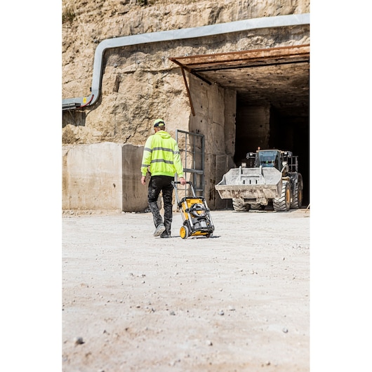 A person wearing a high-visibility jacket is pulling a DEWALT DCMPW1600 portable pressure washer on wheels across a dusty worksite with heavy machinery visible in the background.