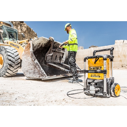 A DEWALT DCMPW1600 pressure washer is being used at a construction site to clean the bucket of a heavy-duty Komatsu loader. The pressure washer is positioned in the foreground with a worker in high-visibility clothing operating it.