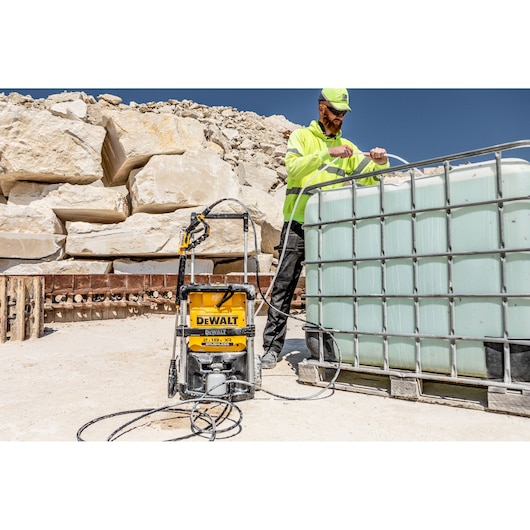 A DEWALT DCMPW1600 pressure washer is positioned outdoors next to a large water tank. A person wearing a high-visibility jacket and cap is handling a hose attached to the tank. The setting features rocky terrain and construction materials.