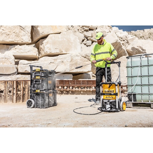 A person wearing a high-visibility jacket and cap is using a DEWALT pressure washer in an outdoor industrial setting to clean stacked black toolboxes. Large stone blocks and a water container are visible in the background.