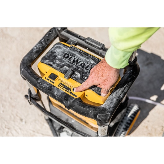 Close-up of a DEWALT portable pressure washer with a person's hand pressing a button on the control panel. The unit is dusty and appears to be used outdoors.