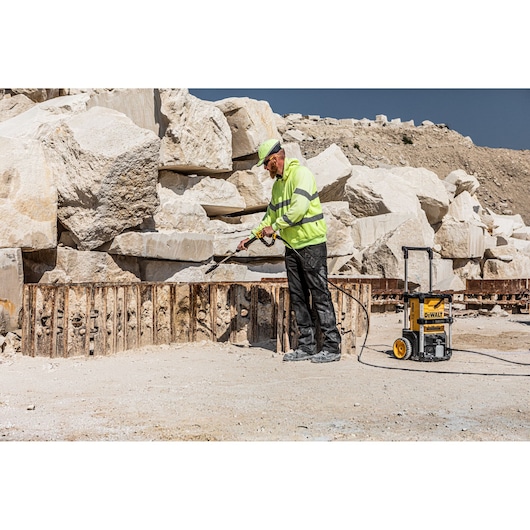 A worker in a high-visibility jacket and helmet uses a DEWALT portable pressure washer (DCMPW1600) outdoors, cleaning a metal surface at a quarry site with large stone blocks in the background.