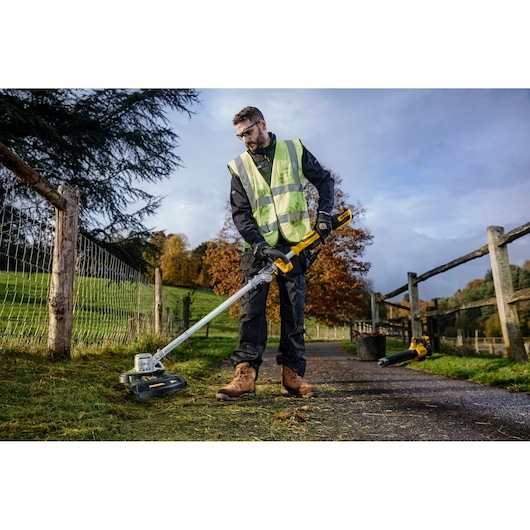 A person wearing a high-visibility vest and work boots is using a DEWALT cordless string trimmer (model DCMST561N) to cut grass along a fence in an outdoor rural setting.