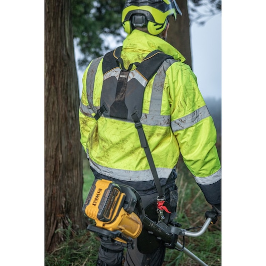Person wearing safety gear, including a helmet and reflective jacket, operating a DEWALT DCMST922N outdoor power tool, likely a grass trimmer, in a wooded outdoor area.