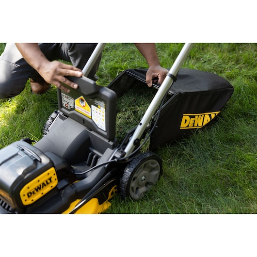 A close-up of a DEWALT cordless lawn mower on grass. A person is emptying the grass collection bag, which is attached to the mower.