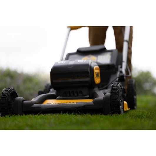 Close-up view of a DEWALT cordless lawn mower in use outdoors, showing the front wheels, mower deck, and battery
