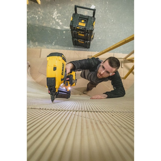 Person using a DEWALT nailer tool on wood surface, with DEWALT storage boxes visible in the background.
