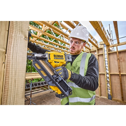 A worker wearing a high-visibility vest and a hard hat is using a DEWALT DCN930N cordless nail gun to fasten wooden beams at a construction site.