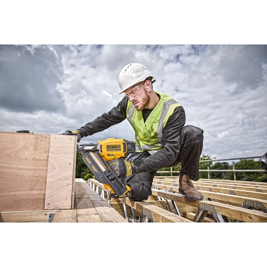 A construction worker wearing a safety helmet and high-visibility vest is using a yellow DEWALT DCN930N nailer to secure wooden boards at an outdoor building site. The worker's face is blurred for privacy.