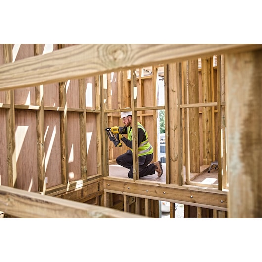 Person wearing a high-visibility vest kneeling inside a wooden framed construction site, operating a DEWALT nail gun.