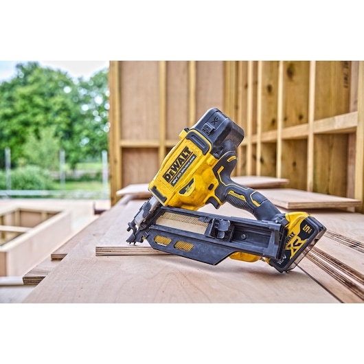 A DEWALT cordless nailer tool, model DCN930N, placed on a stack of wooden boards at a construction site with wooden framing and greenery in the background.