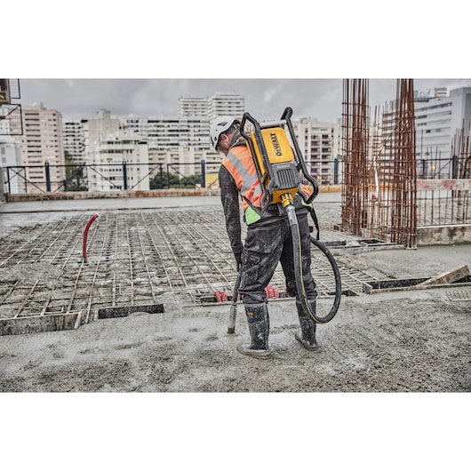 A construction worker wearing a safety helmet, high-visibility vest, and boots is using a DEWALT concrete vibrator on a large outdoor building site. The worker has the DEWALT device strapped to their back, and is standing on wet concrete near metal rebar structures, with city buildings visible in the background.