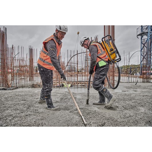 Two construction workers wearing safety helmets and orange reflective vests are working on a concrete slab at an outdoor construction site. One worker is using a DEWALT concrete vibrator backpack, while the other is spreading fresh concrete with a rake. Exposed rebar and industrial equipment are visible in the background.