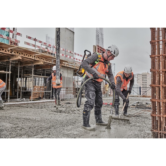 Construction workers wearing safety gear, including orange vests and helmets, use tools to spread and smooth concrete on a building site.